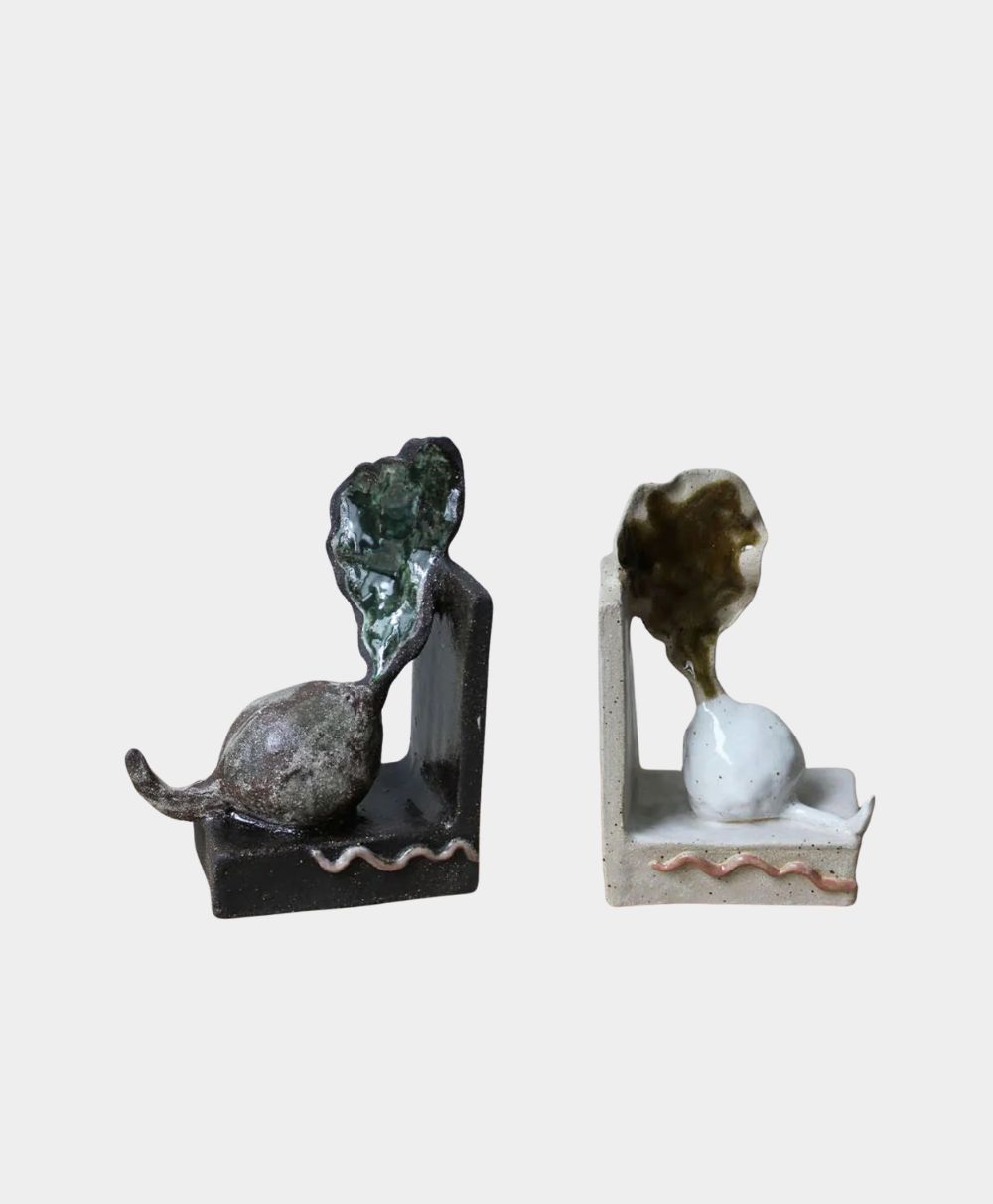 Two abstract ceramic sculptures, each featuring a rounded base with a tail-like extension, topped with an irregular, textured form, set against a plain white background. One sculpture is dark, the other is light.