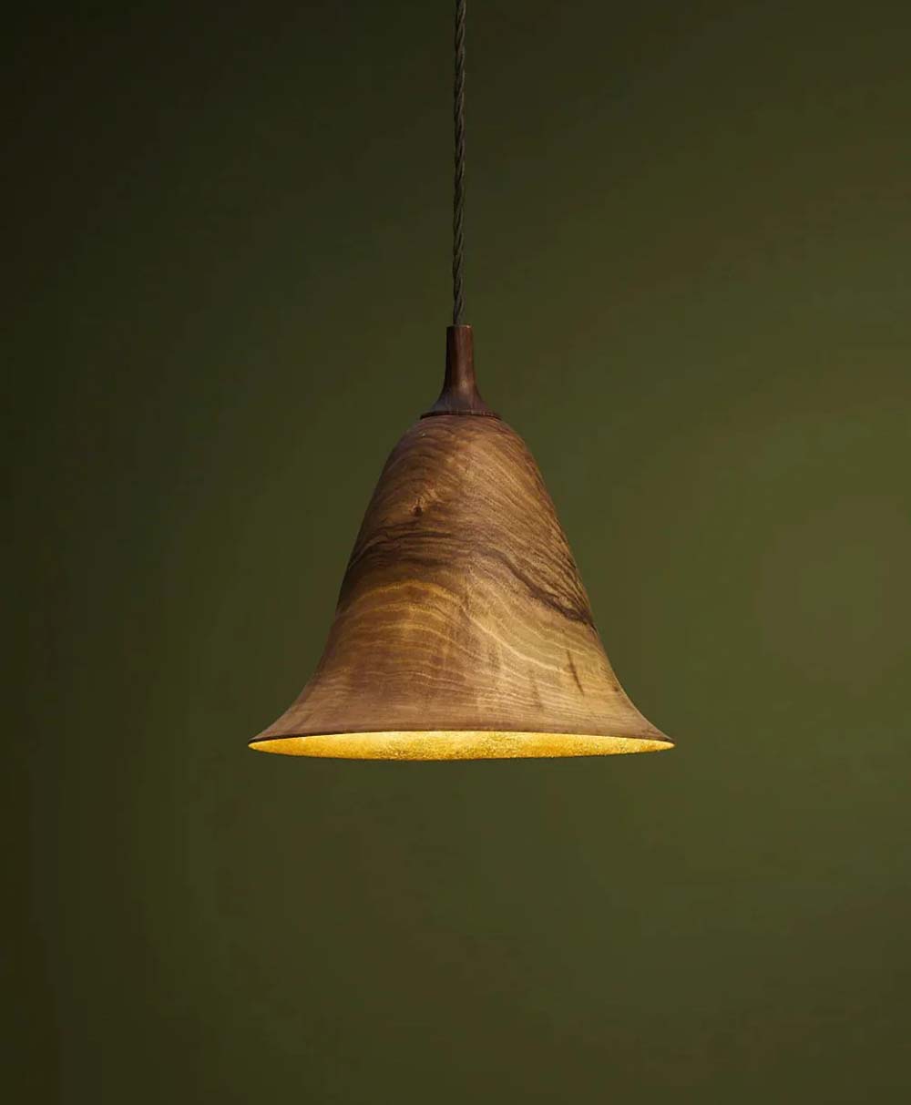 A wooden pendant light with a flared, bell-shaped shade hangs from a twisted black cord against an olive green background. The light glows warmly, highlighting the wood grain.