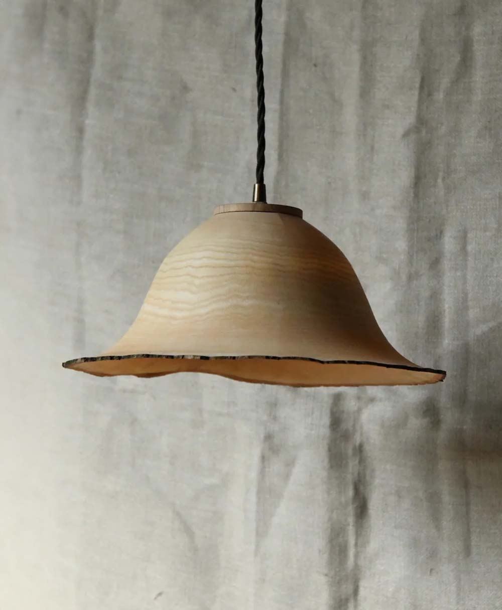 A handcrafted wooden pendant lamp with a wavy, natural edge hangs from a twisted black cord against a textured, light-colored fabric background.
