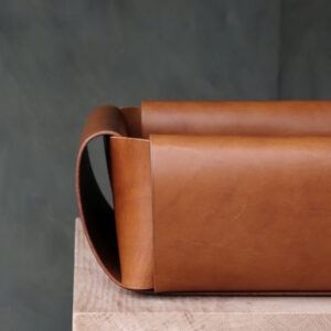 A close-up of a rectangular, brown leather tray with curved side handles, placed on top of a light wooden surface against a dark, blurred background.