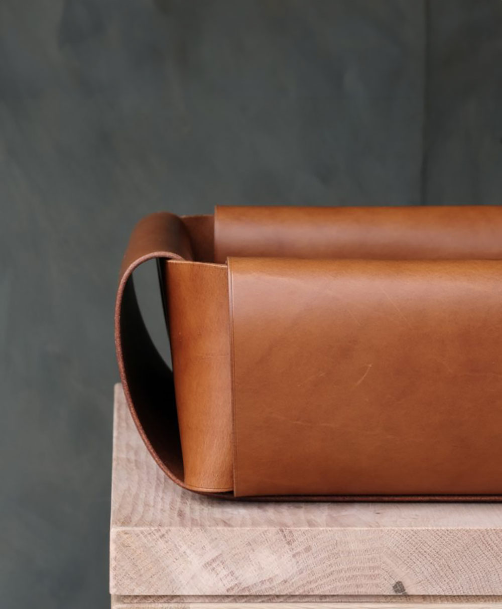 A close-up of a rectangular, brown leather tray with curved side handles, placed on top of a light wooden surface against a dark, blurred background.