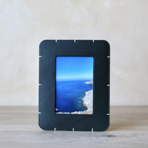 A black picture frame with white stitching sits on a light wooden surface, displaying a photograph of a rocky coastline with deep blue water and a pale blue sky in the background.