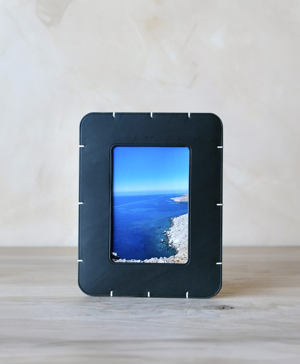 A black picture frame with white stitching sits on a light wooden surface, displaying a photograph of a rocky coastline with deep blue water and a pale blue sky in the background.