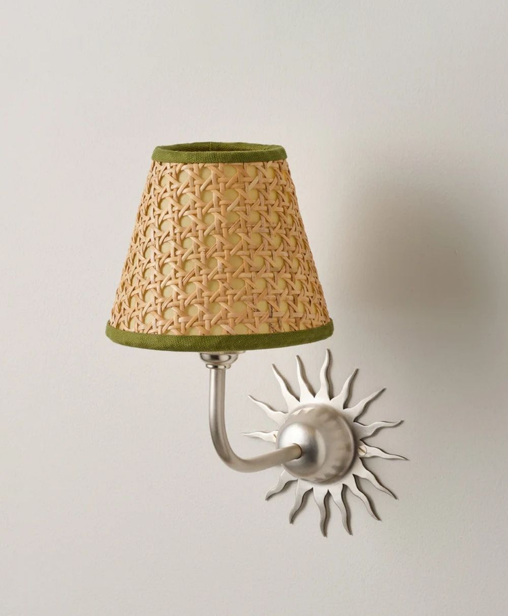 A wall-mounted sconce lamp with a woven rattan shade, green trim, and a brushed metal base shaped like a sunburst, attached to a white wall.