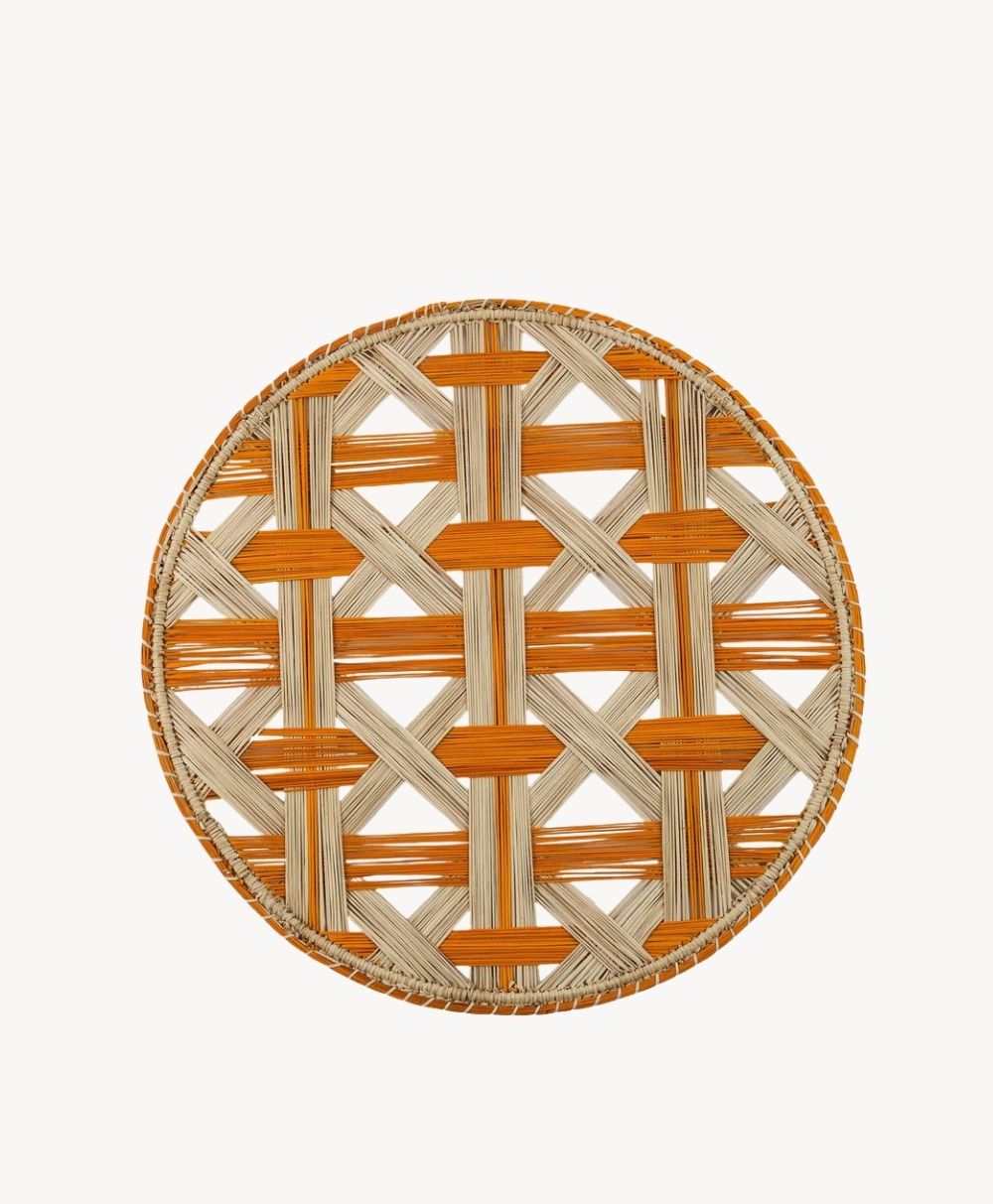 A round, flat wicker basket with a crisscross woven pattern in natural beige and orange hues. The basket has a sturdy rim, also accented with orange weaving.