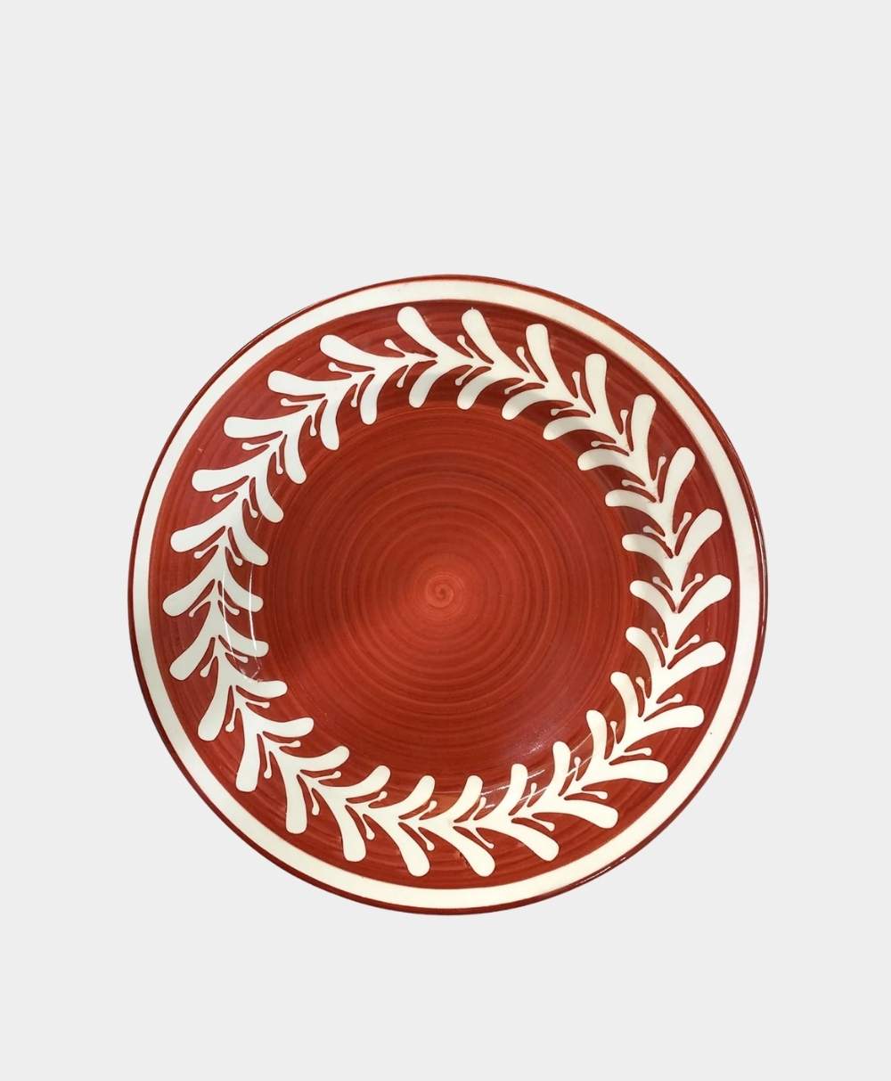 A round ceramic plate with a red background and a white leafy vine pattern circling the rim, viewed from above on a plain light surface.
