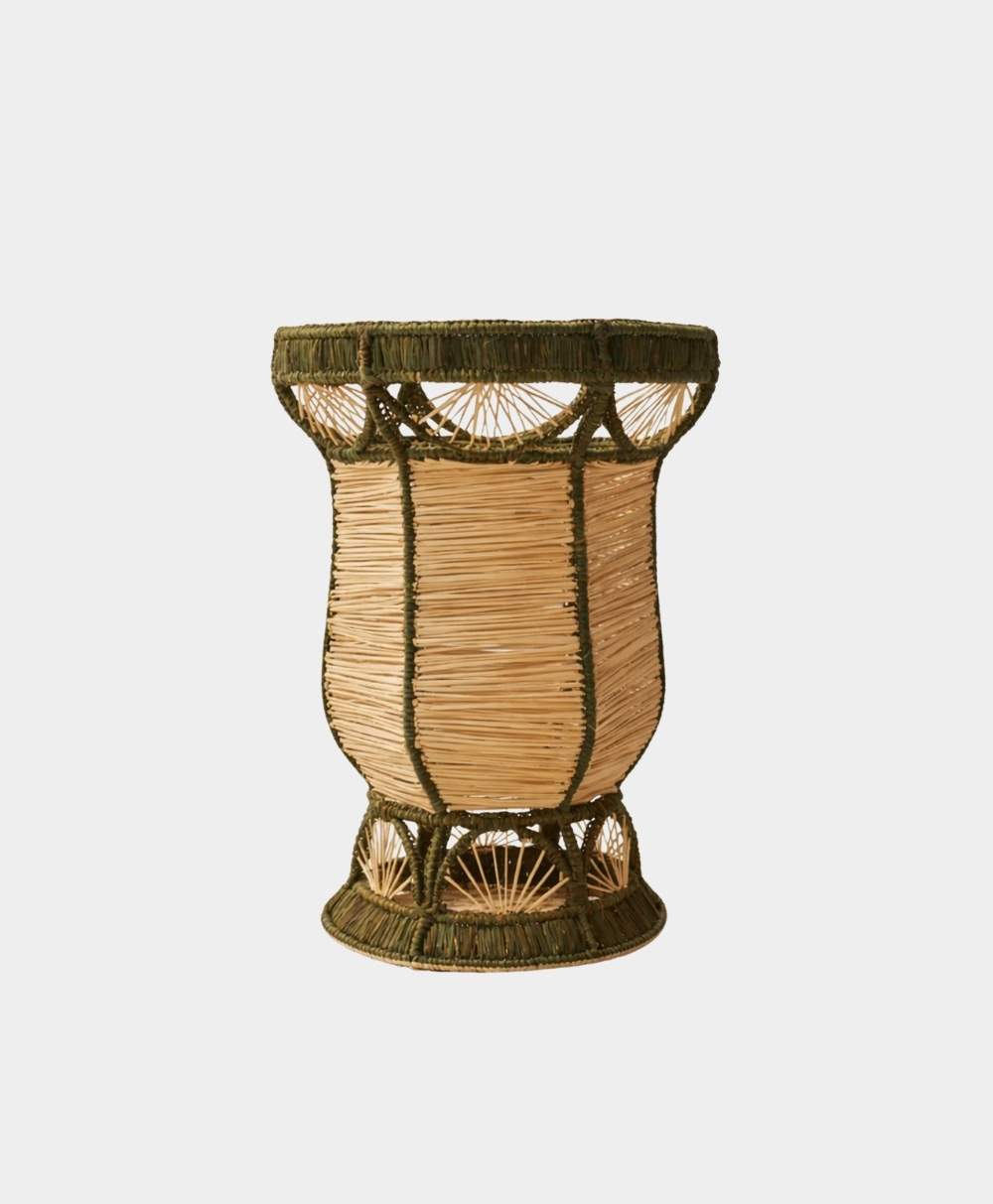 A handcrafted woven basket with an hourglass shape, featuring natural-colored fibers and green accents around the rim, base, and vertical supports, set against a plain white background.