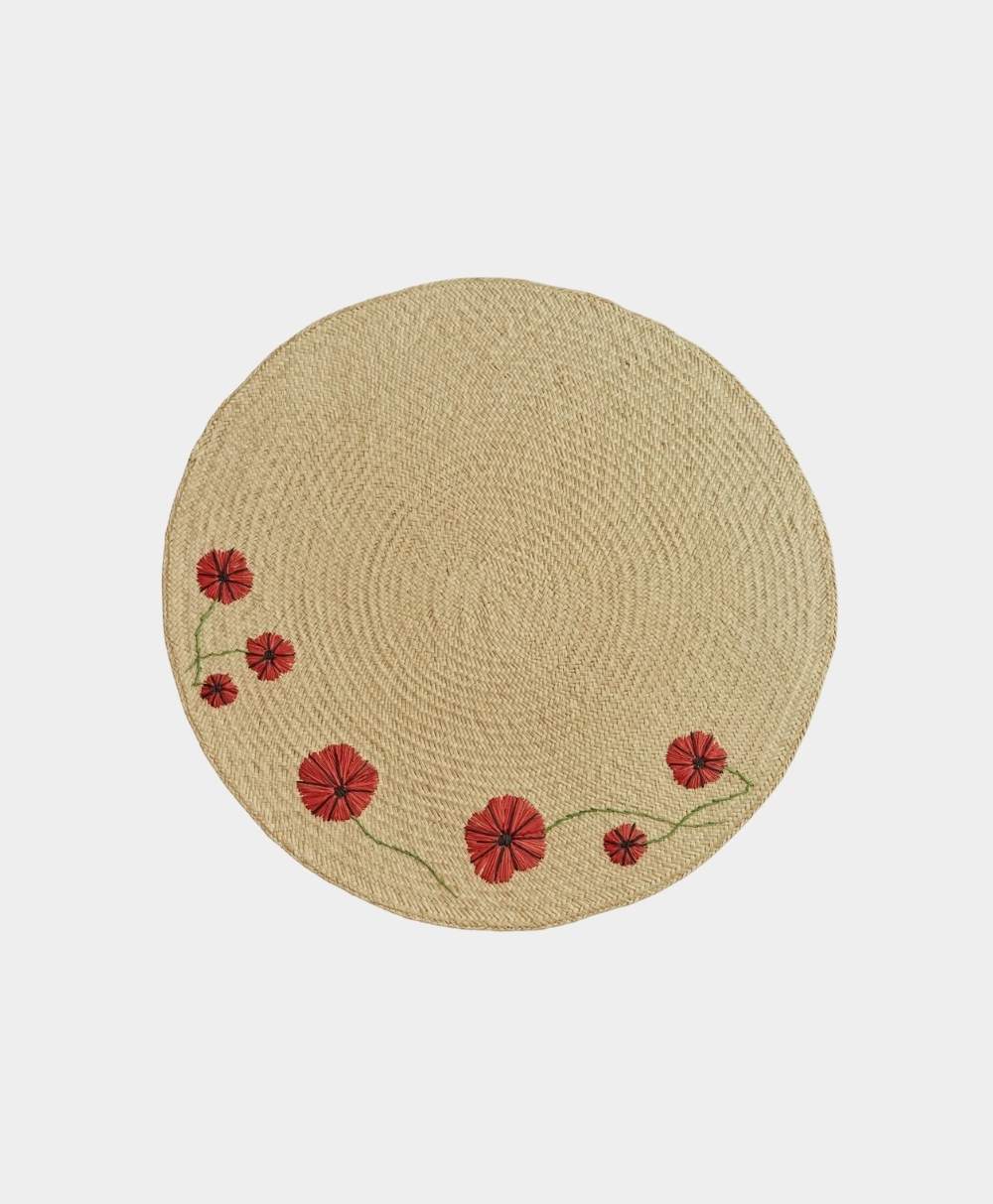A round, woven straw placemat with embroidered red flowers and green stems arranged along one edge, set against a plain white background.