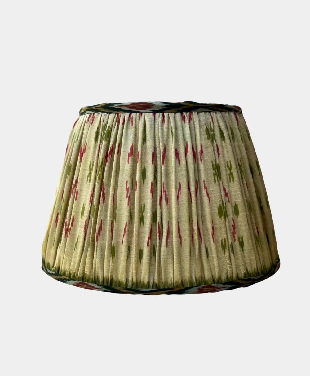 A pleated fabric lampshade with a cream background and green and red abstract patterns, set against a plain light gray backdrop.