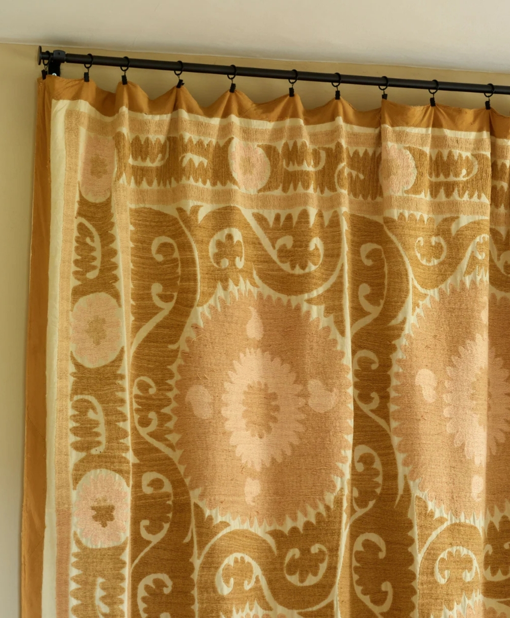 A beige and gold curtain with a decorative, patterned design featuring circular and floral motifs hangs from a black curtain rod with rings.