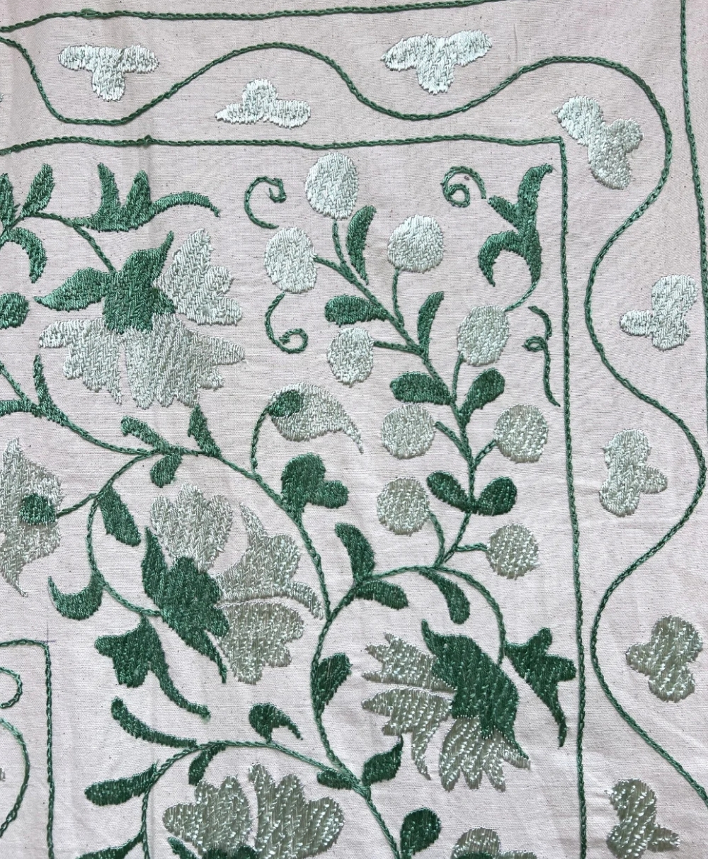 Embroidered fabric with a floral and leafy pattern in shades of green and silver on a light background. The design features vines, flowers, and decorative border elements.