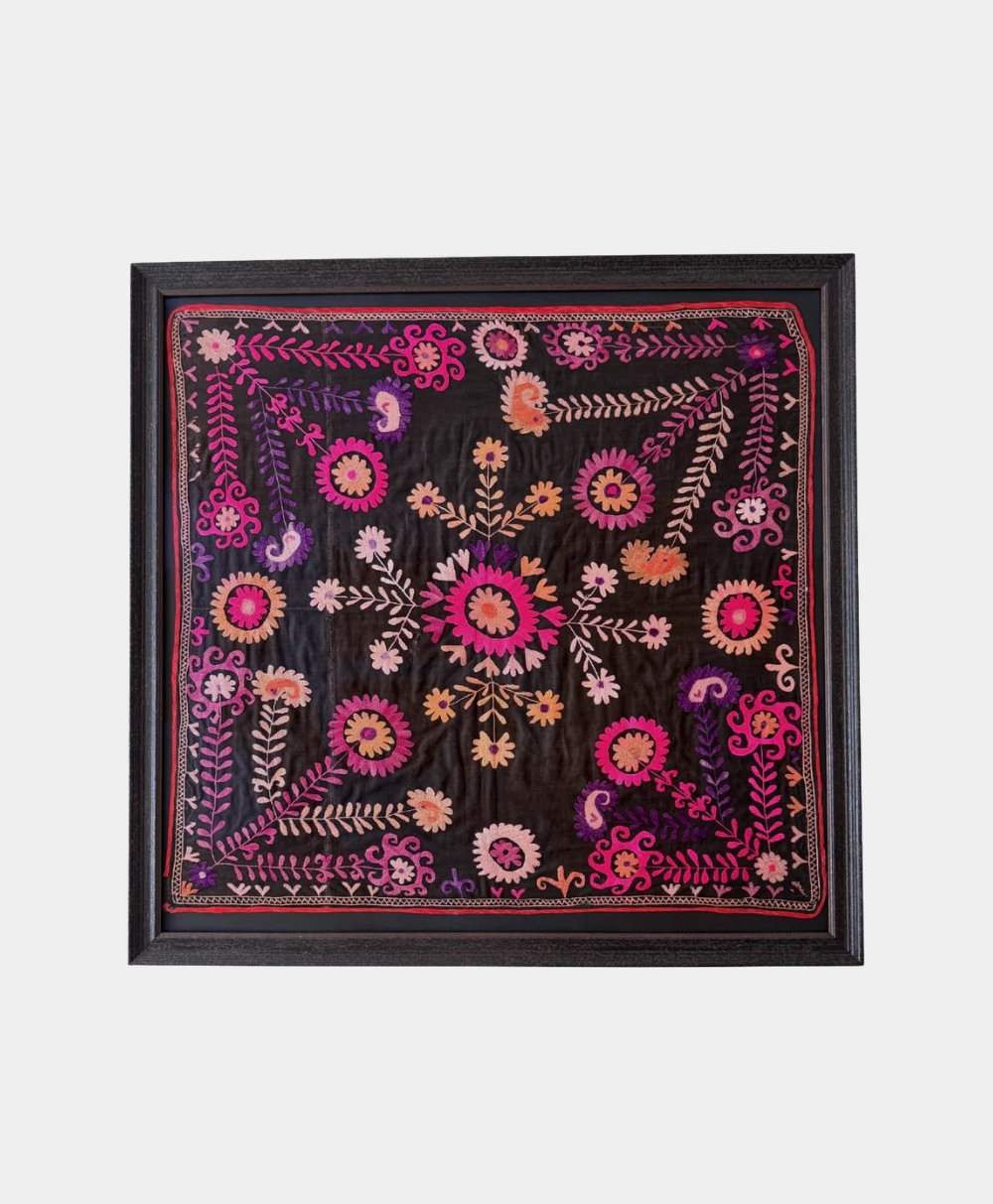 A framed textile with intricate floral and leaf embroidery in pink, orange, purple, and beige threads on a black background, featuring symmetrical patterns and bold colors.
