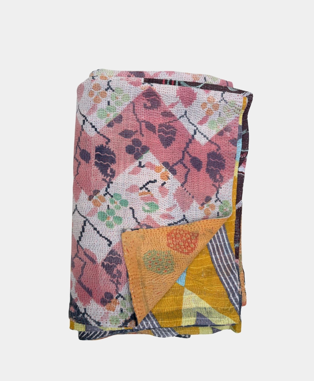 A folded quilt with a pink, white, and black floral and geometric pattern on top, revealing a corner with yellow, orange, blue, and striped designs.