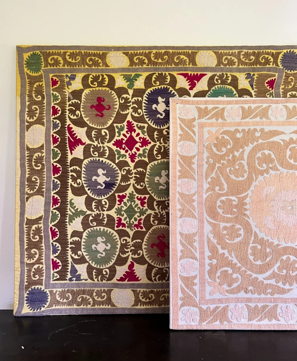 Two embroidered rugs with ornate patterns are displayed upright. The larger rug has a colorful, intricate design with red, gold, green, and purple details, while the smaller rug has a subtler beige and white floral pattern.