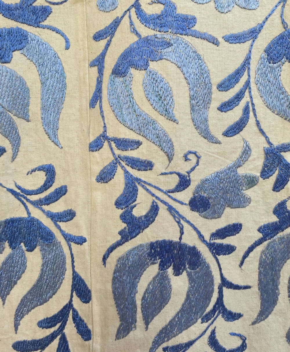 Close-up of fabric with an embroidered floral and leaf pattern in various shades of blue on a light beige background. The design features intricate, swirling vines and stylized flowers.