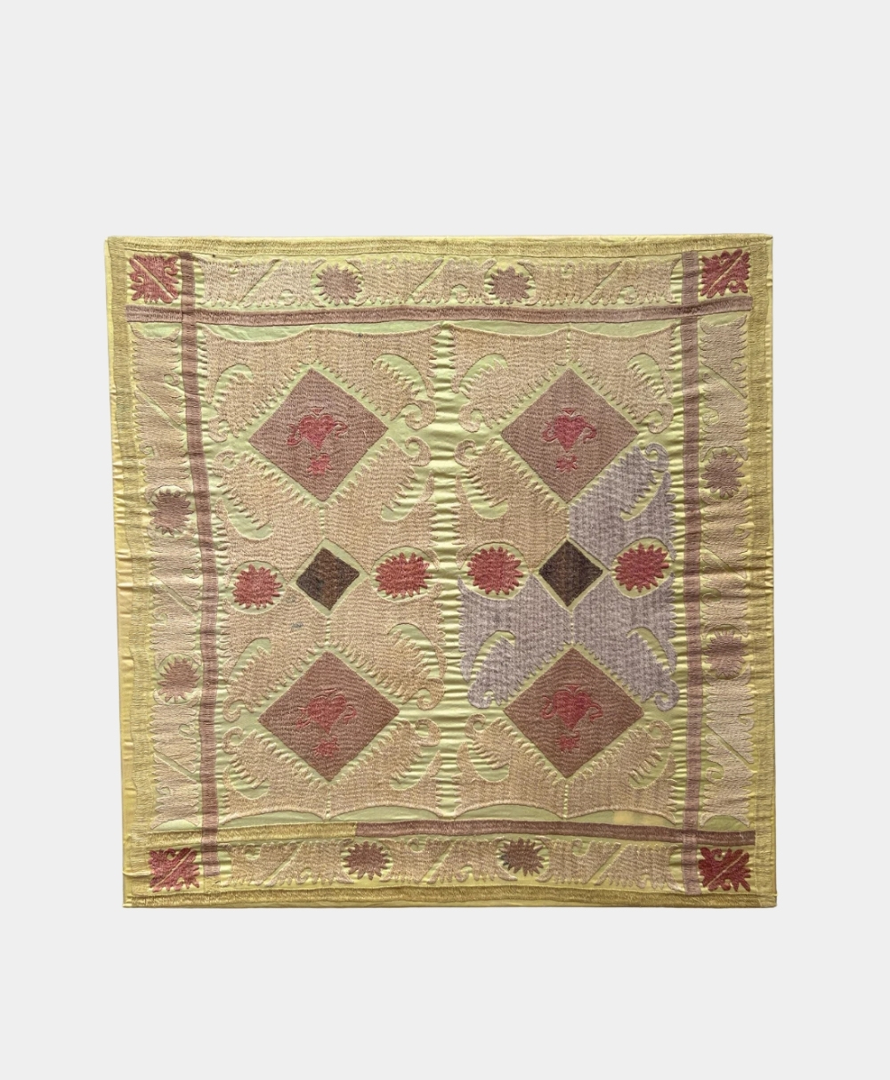 A square quilt with a geometric pattern featuring diamonds, floral motifs, and swirls in muted tones of beige, red, green, and brown, bordered by a yellow and red design.