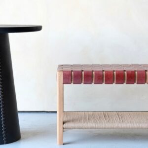 A modern interior features a round black table with stitched detailing next to a wooden bench with a woven reddish-brown leather seat and a woven natural fiber shelf, set against a light-colored wall and pale floor.