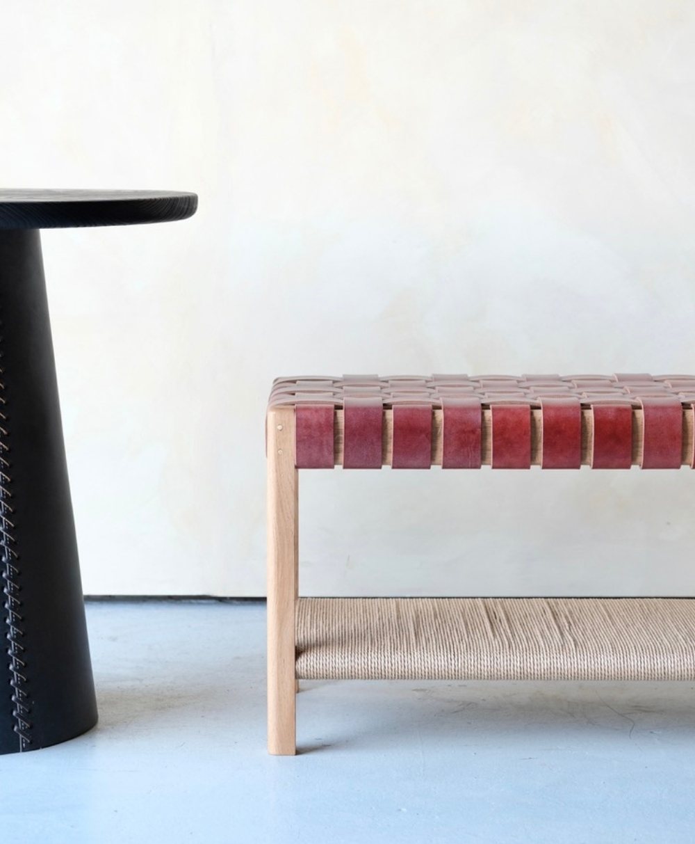 A modern interior features a round black table with stitched detailing next to a wooden bench with a woven reddish-brown leather seat and a woven natural fiber shelf, set against a light-colored wall and pale floor.