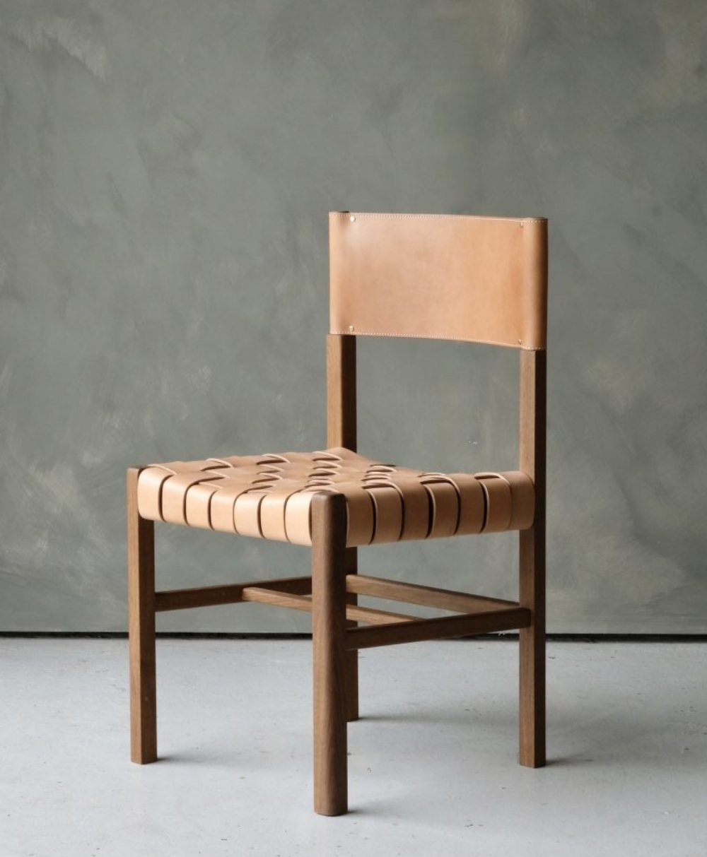 A minimalist chair with a wooden frame, featuring a woven tan leather seat and a matching leather backrest, set against a plain gray textured wall.
