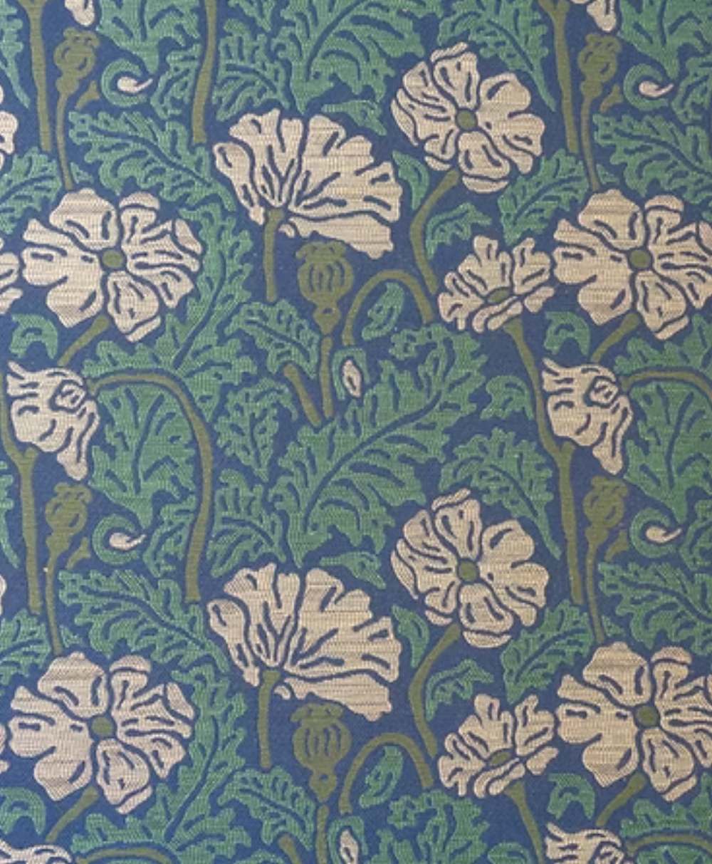 A fabric pattern featuring white flowers with green stems and leaves on a blue background, arranged in a repeating, intricate design reminiscent of vintage or Art Nouveau styles.