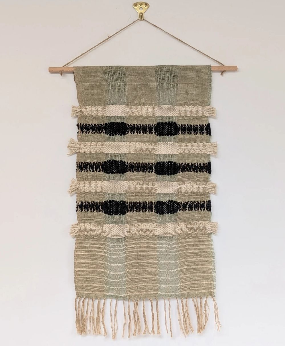 A handwoven wall hanging features beige and black geometric patterns, horizontal stripes, and fringed edges, displayed on a wooden rod against a light-colored wall.