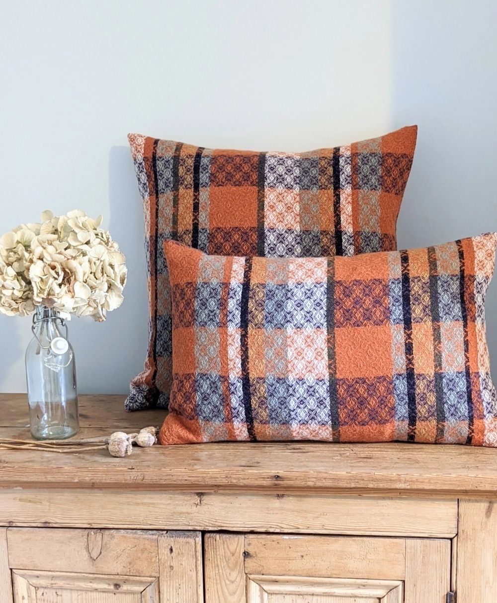 Two rectangular pillows with orange, blue, and cream plaid patchwork patterns rest on a wooden cabinet. A glass vase with white dried flowers sits beside them against a light-colored wall.