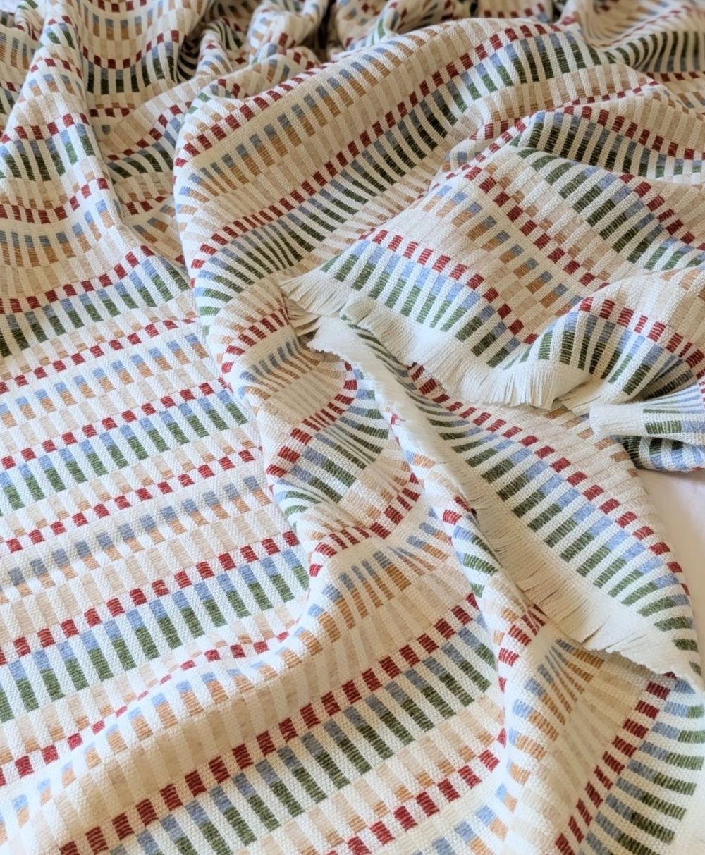 A close-up of a textured fabric featuring horizontal rows of small, colorful rectangles in red, green, blue, and beige on a light background. The fabric appears softly wrinkled.