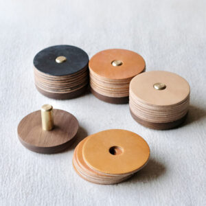 Five stacks of round leather coasters, each with a hole in the center and a circular wooden base with a brass rod. The leather coasters are in shades of tan, brown, and black, displayed on a light textured surface.