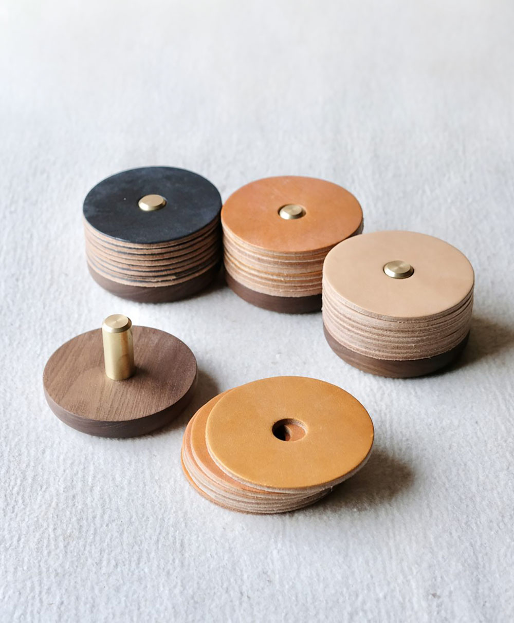 Five stacks of round leather coasters, each with a hole in the center and a circular wooden base with a brass rod. The leather coasters are in shades of tan, brown, and black, displayed on a light textured surface.