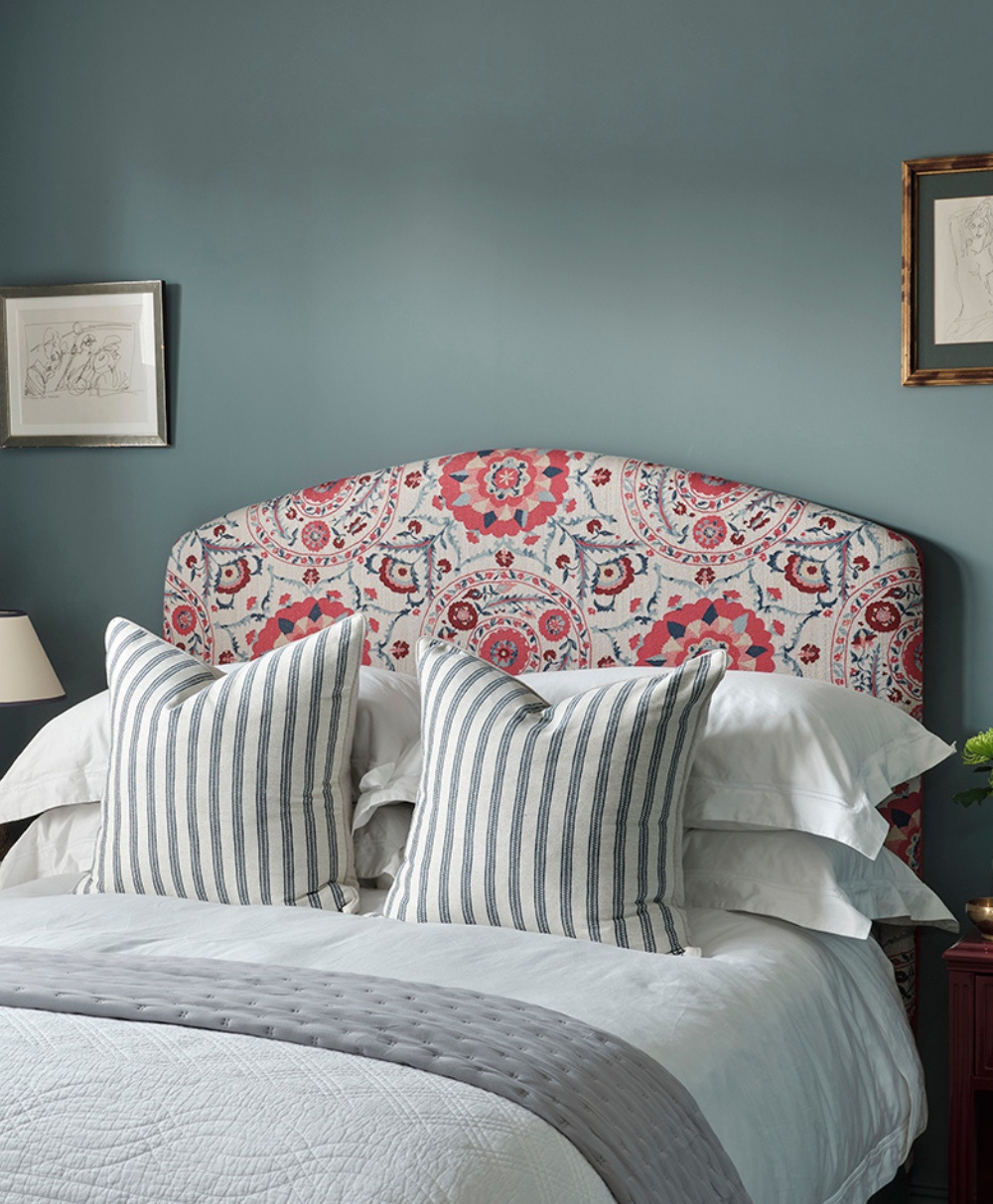 A neatly made bed with striped pillows and a quilted blanket sits against a blue wall. The headboard is upholstered with a floral print in pink, red, and white. Two framed pictures hang on the wall above the bed.
