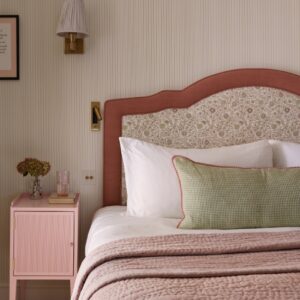 A cozy bedroom features a bed with a patterned headboard, white and green pillows, pink quilted bedding, and a pink nightstand with books, a glass, and dried flowers, next to a wall sconce.