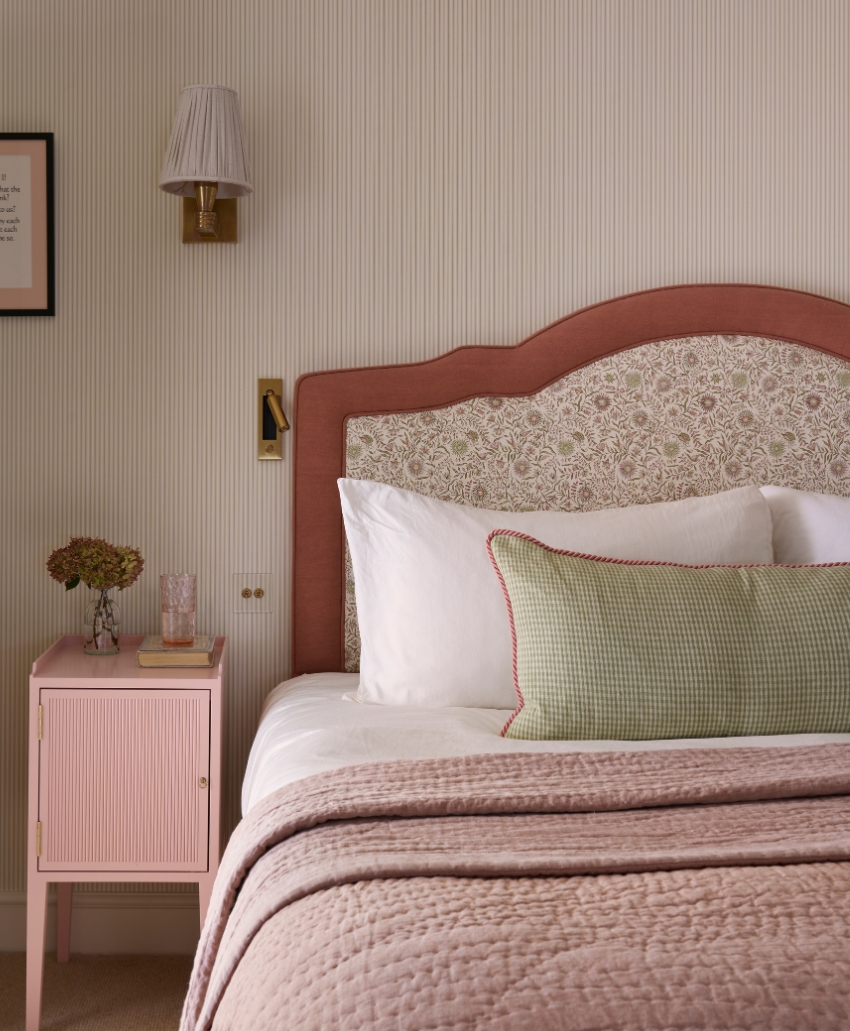 A cozy bedroom features a bed with a patterned headboard, white and green pillows, pink quilted bedding, and a pink nightstand with books, a glass, and dried flowers, next to a wall sconce.
