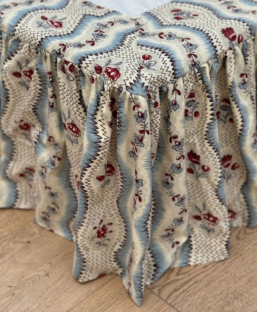A table covered with a draped fabric featuring a wavy blue, white, and cream pattern with red floral accents, set on a wooden floor.