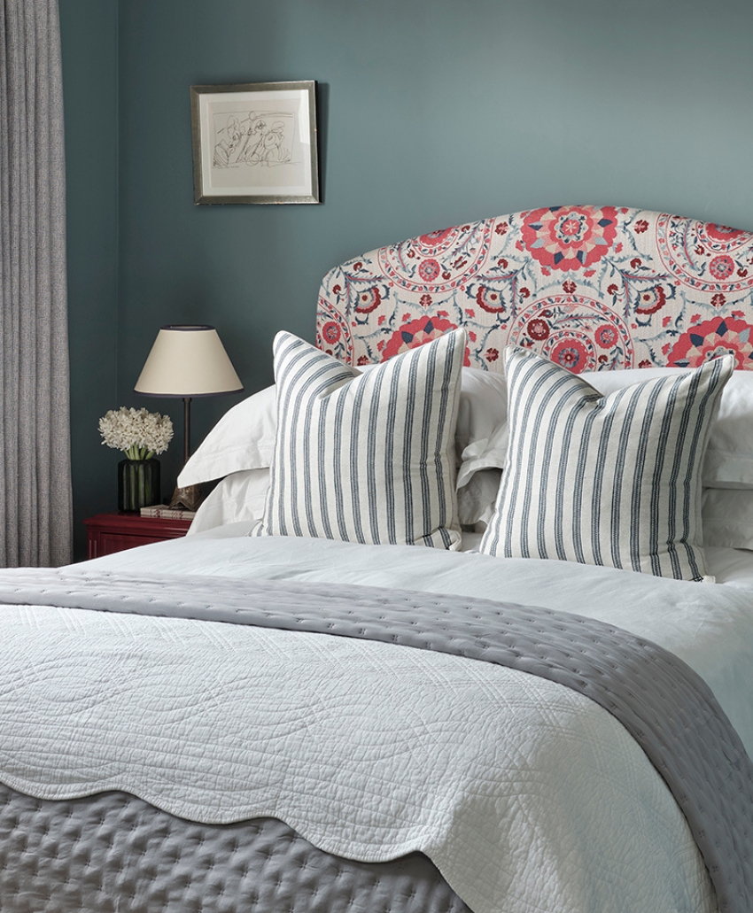 A cozy bedroom with a bed featuring a floral-patterned headboard, striped pillows, and layered grey and white bedding. A bedside table holds a lamp and flowers, and a framed picture hangs on the blue wall.
