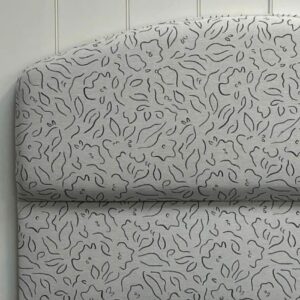 A padded headboard with a curved top, upholstered in light fabric featuring an abstract floral or leaf pattern in dark lines, set against a white paneled wall.