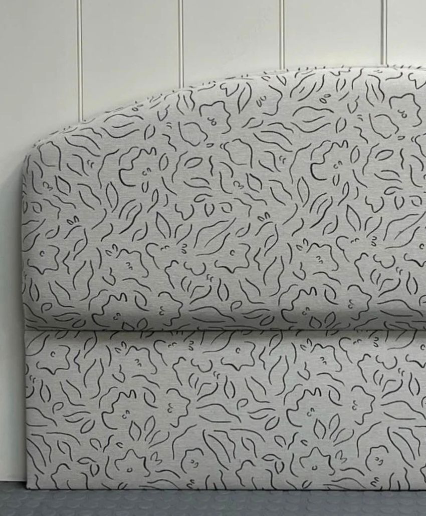 A padded headboard with a curved top, upholstered in light fabric featuring an abstract floral or leaf pattern in dark lines, set against a white paneled wall.