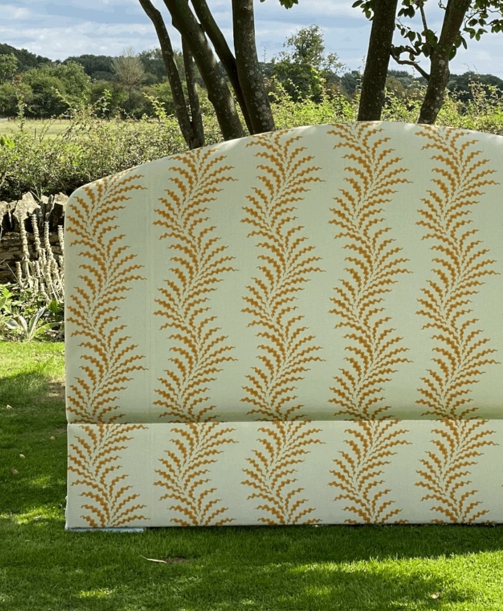 A patterned upholstered headboard with a yellow and orange leaf-like design stands on a green lawn with trees, stone wall, and countryside visible in the background.