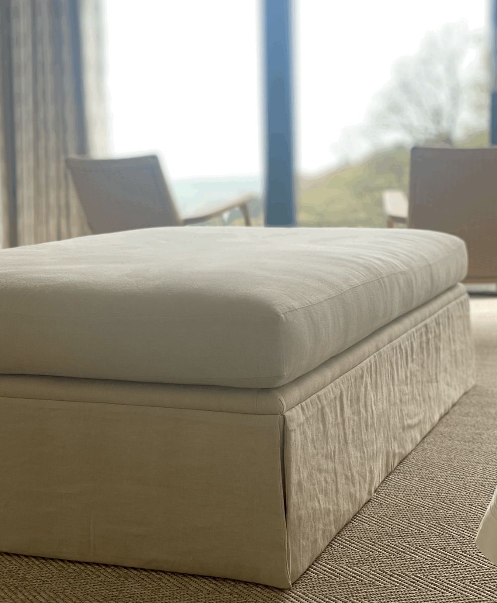 A close-up of a beige upholstered bench or ottoman in a bright room with soft natural light, two chairs, and large windows in the background overlooking greenery.