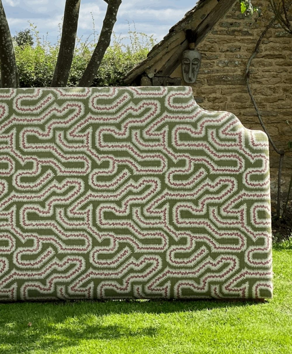 A patterned upholstered headboard with green, white, and red zigzag designs stands on a grassy lawn, with trees and a stone building partially visible in the background.