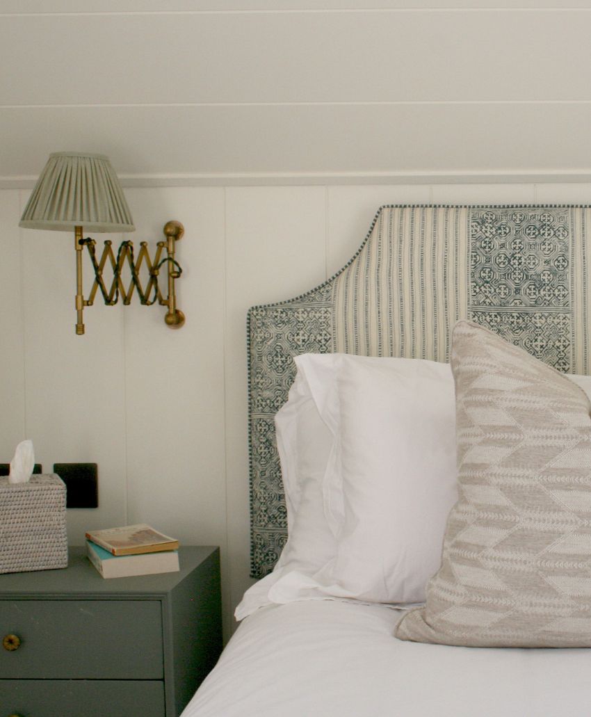 A cozy bedroom corner with a green nightstand holding a tissue box and a book, a bed with white linens and a patterned pillow, and a wall-mounted lamp above the nightstand.