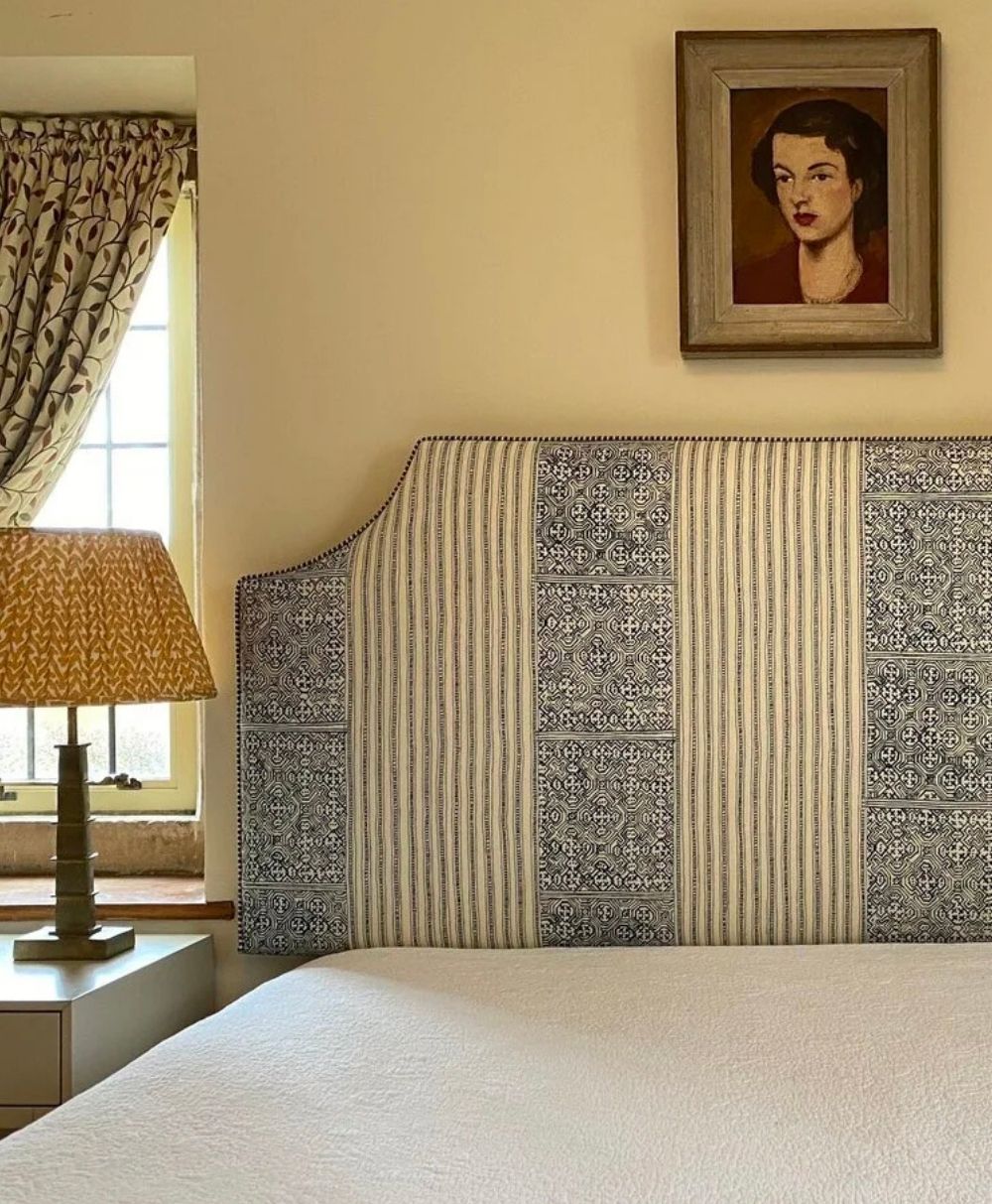 A bedroom with a patterned headboard, a white bed, a nightstand with a yellow patterned lamp, floral curtains, and a framed portrait of a woman on the wall above the bed.