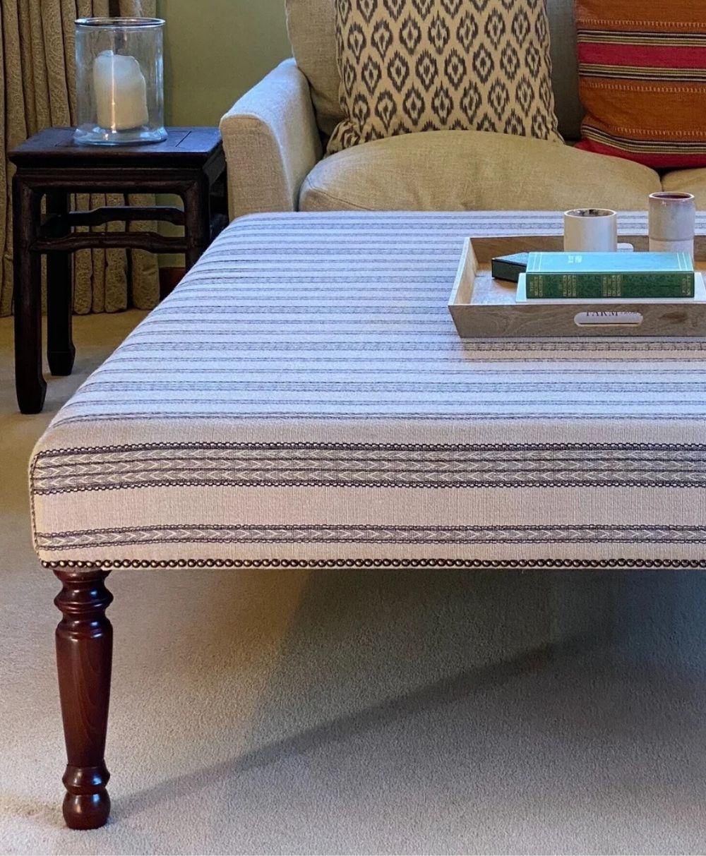 A striped upholstered ottoman with dark wooden legs sits on a beige carpet. A tray with books and a candle is on top. In the background, a beige sofa with patterned pillows and a small side table are visible.