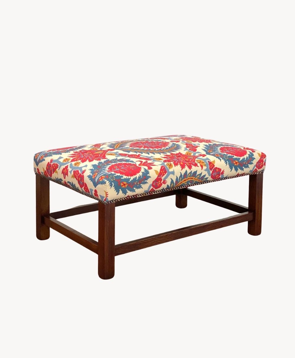 Rectangular wooden ottoman with a cushioned top, upholstered in a vibrant fabric featuring red, blue, and yellow floral patterns. The base has straight wooden legs and crossbar supports.