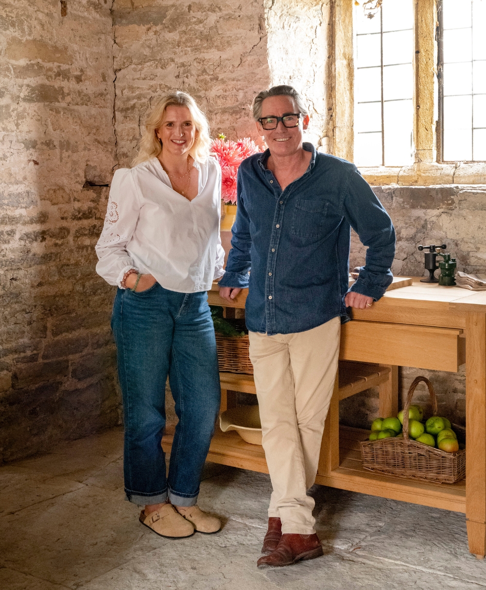 A woman and a man stand smiling in a rustic stone kitchen with a wooden counter, baskets of green apples, and sunlight streaming through a window behind them.