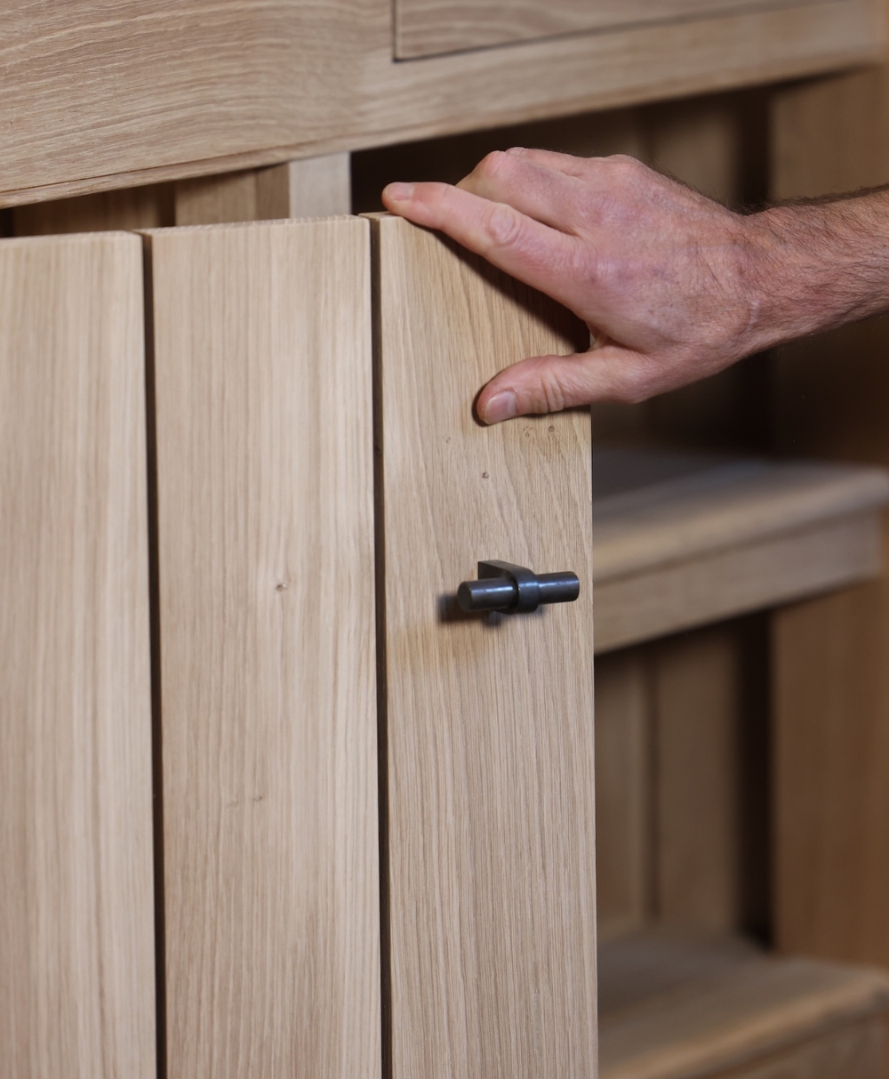 A hand is opening a light wooden cabinet door with a small, dark metal latch. The shelves inside the cabinet are visible.