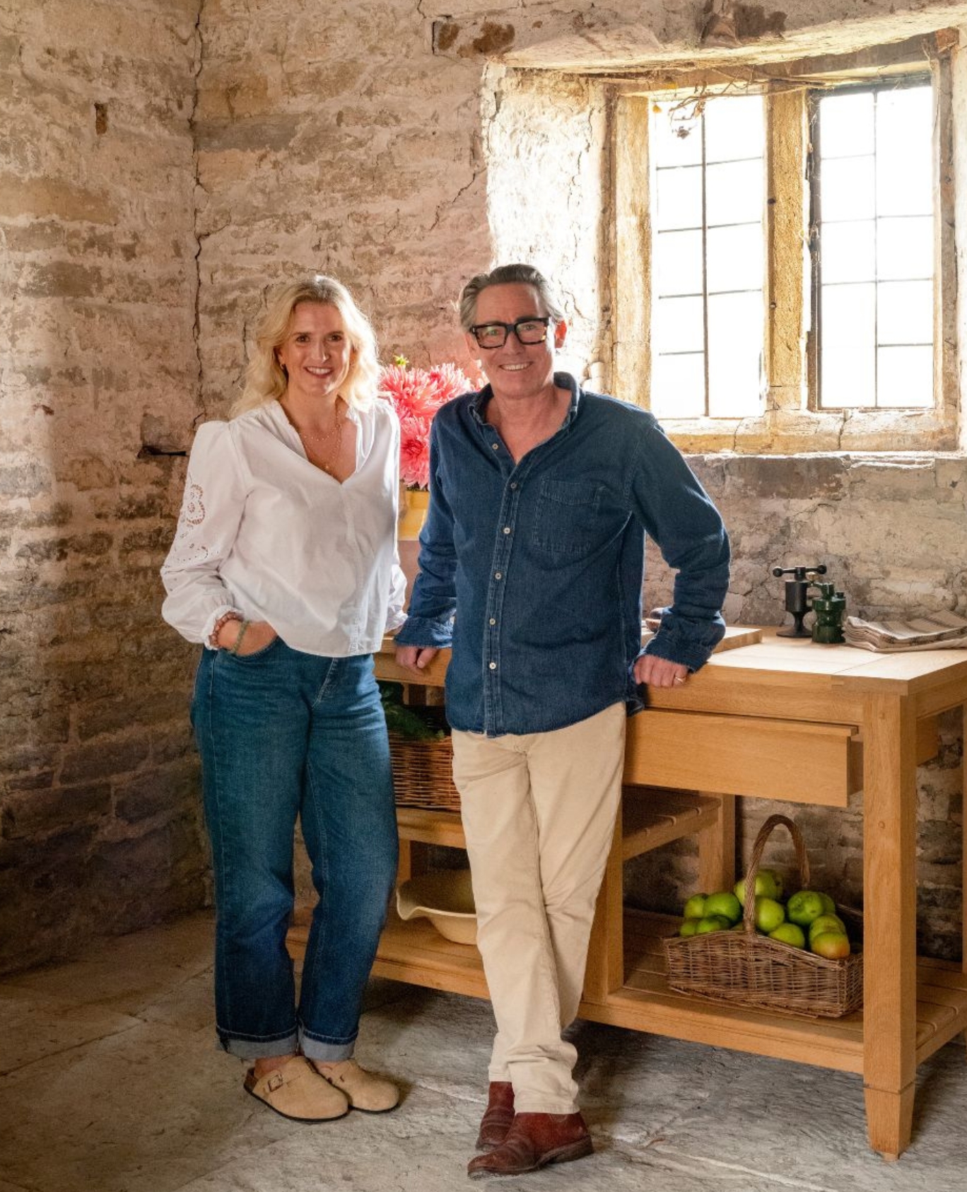 A woman and a man stand smiling in a rustic kitchen with stone walls, a wooden table holding apples and baskets, and sunlight streaming through a small window.