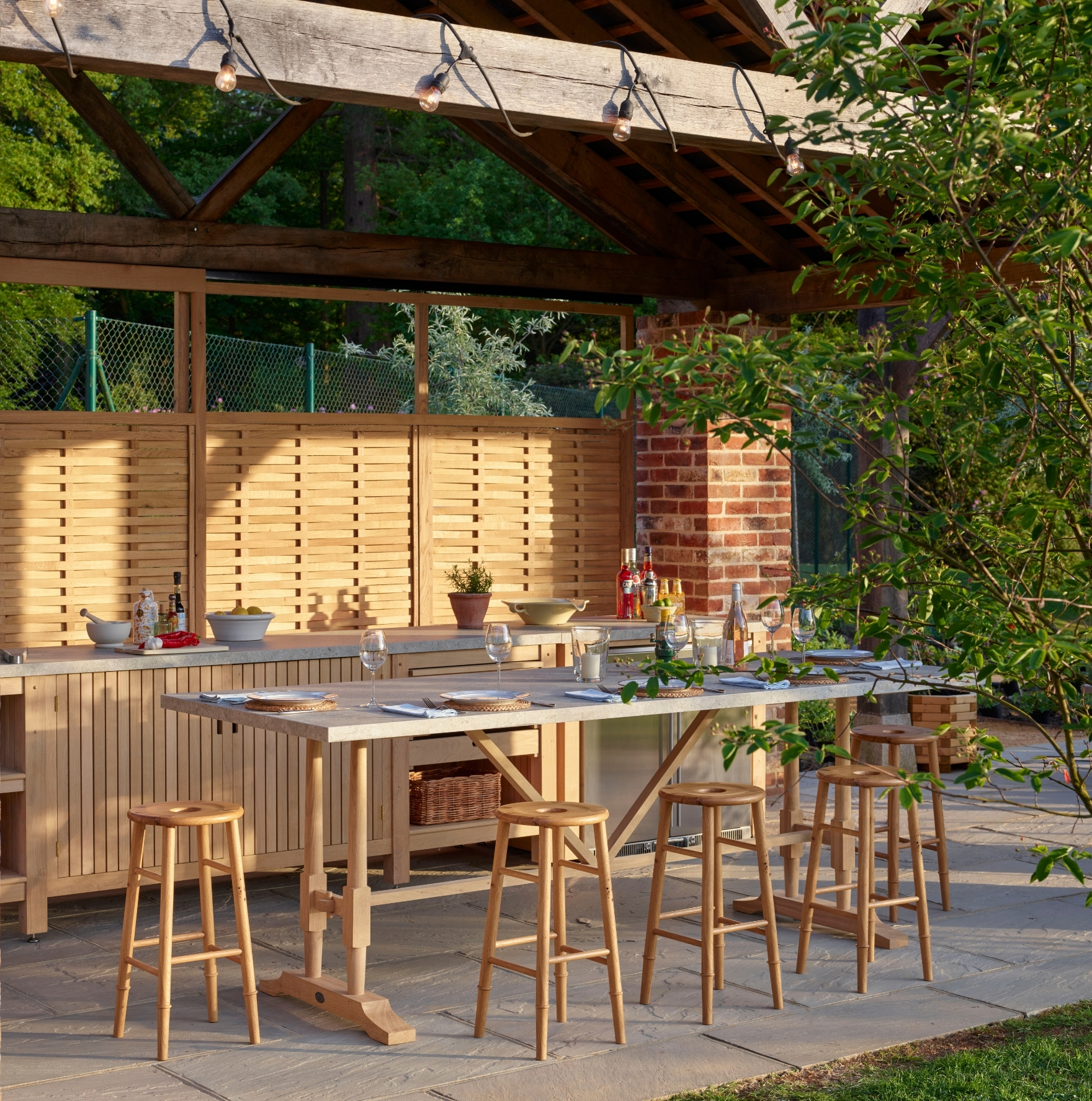A stylish outdoor kitchen and dining area with a wooden table, bar stools, countertop, and shelves, surrounded by greenery and sheltered by a wooden pergola with string lights.