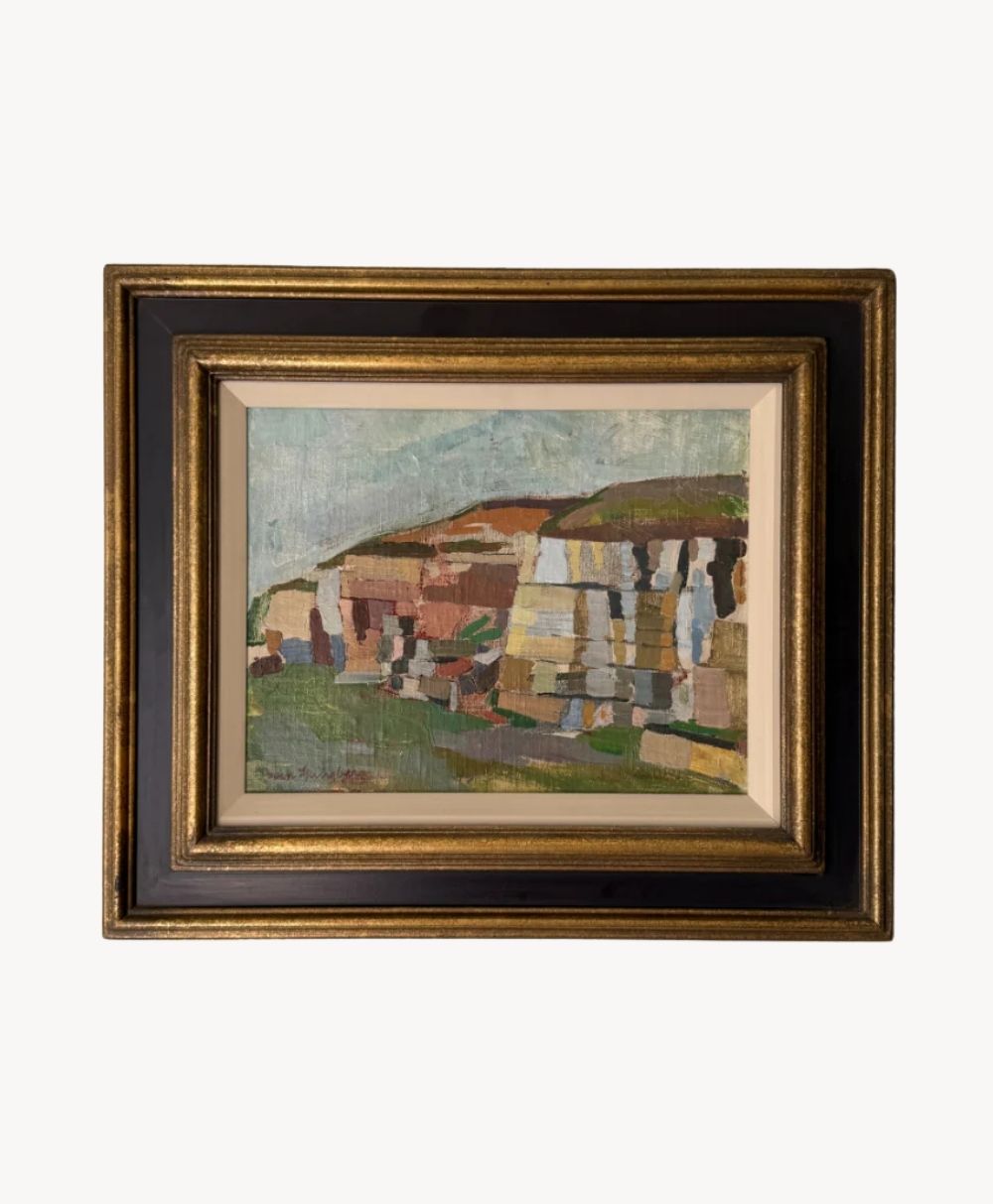 Framed painting depicting a cliffside or rock formation with earthy tones of brown, beige, and green, set against a muted sky. The frame is dark with a gold inner and outer edge.