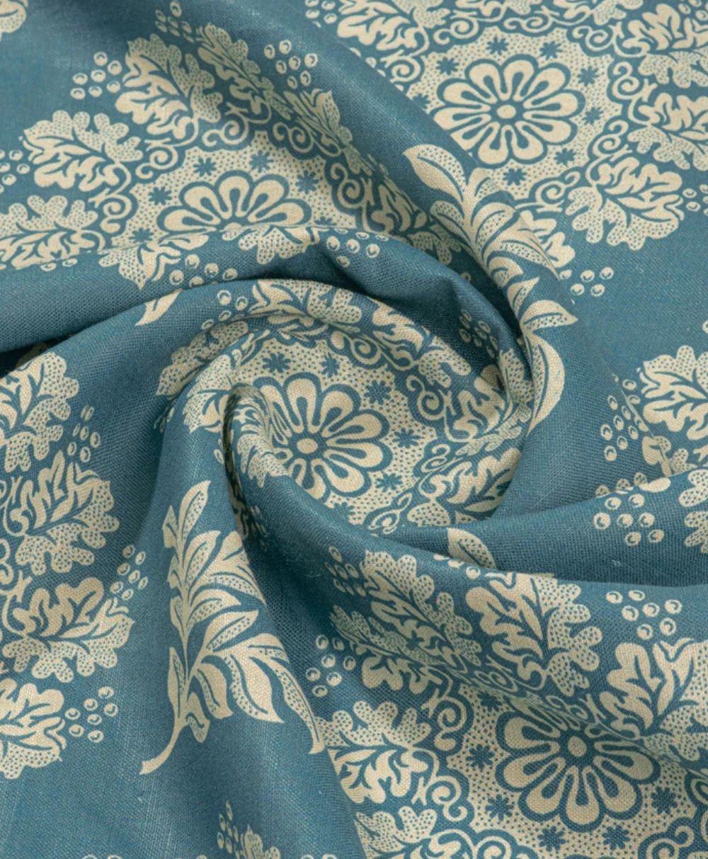 A close-up of blue fabric with intricate cream floral and leaf patterns, slightly gathered in the center to form gentle folds and a spiral-like swirl.