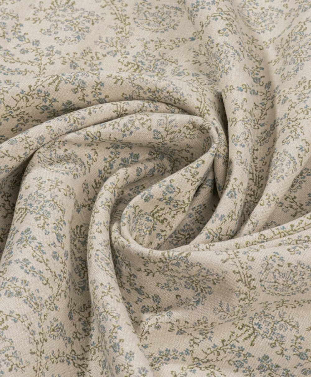A close-up of beige fabric with a delicate, light blue and green floral pattern, softly draped and forming gentle folds and swirls.